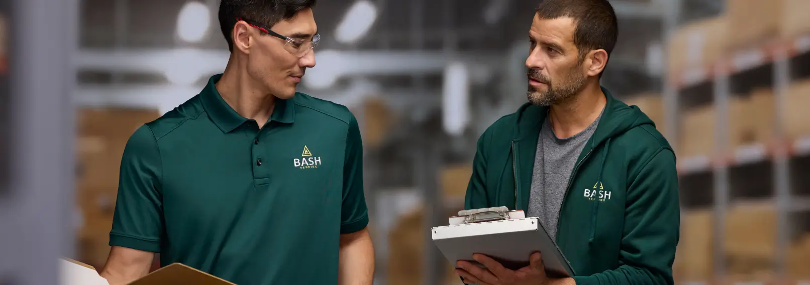 Employees in branded green polo shirt and hoodie reviewing orders in a warehouse