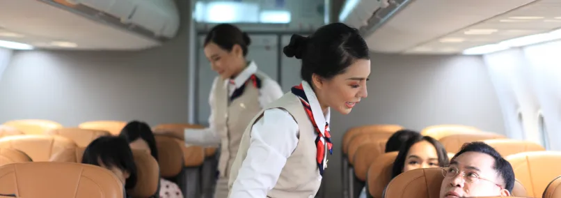 Female flight attendants in cream vests and neck scarves assisting passengers onboard
