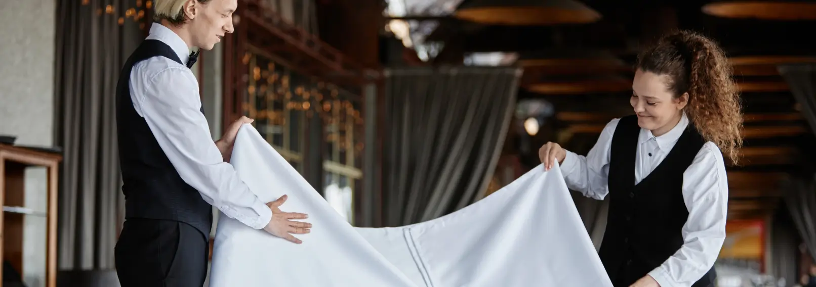 Two hotel workers wearing linen uniforms fold a tablecloth.