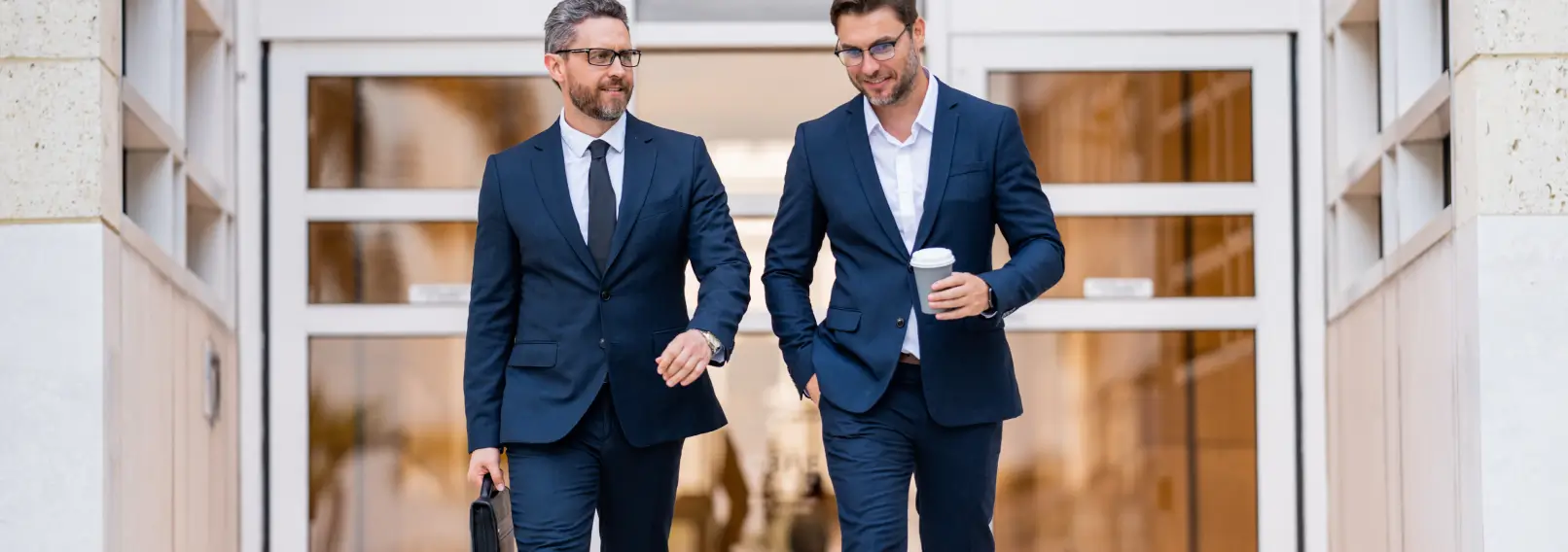 Two businessmen walking out of a building wearing suits.