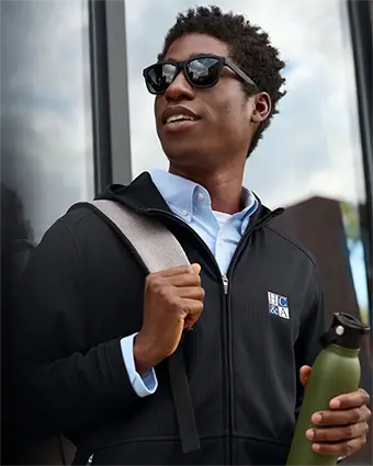 A man wearing sunglasses holds a bag over his shoulder in one hand and a water bottle in the other.